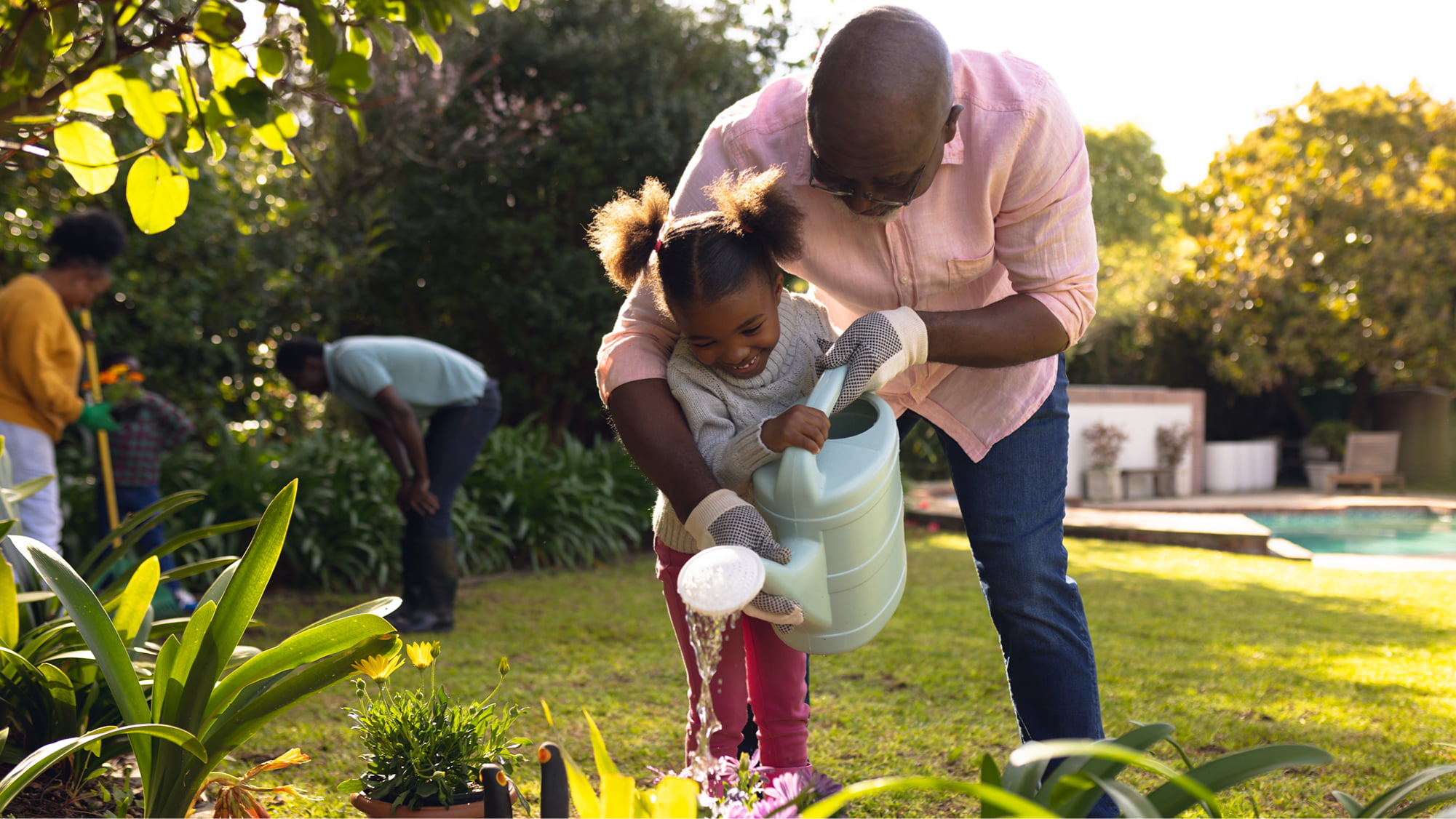 A man and a young girl watering plants together in a sunny garden.